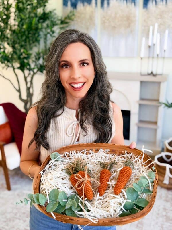 A woman with long gray hair smiles while holding a basket featuring the Mini Crochet Carrot Pattern and decorative greenery, standing in a bright, stylish living room.