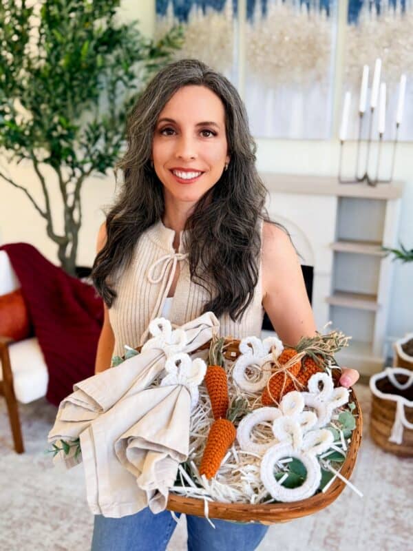A woman with long gray hair holds a basket containing cloth napkins, crocheted bunny decorations, and carrots made using a Mini Crochet Carrot Pattern in a cozy living room.