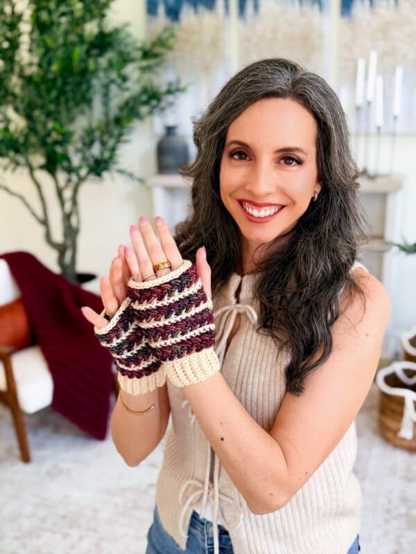 Woman smiling and displaying Wheat Stitch fingerless mittens crochet pattern gloves in cream and maroon colors; indoor setting with plants and decor in the background.