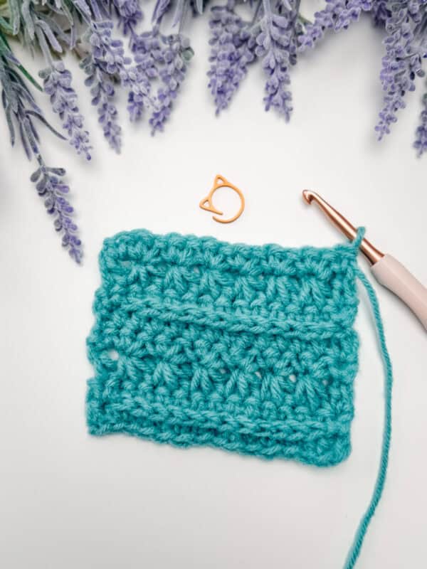 A teal crocheted swatch, a crochet hook, and an orange stitch marker are arranged on a white surface with lavender flowers in the background.