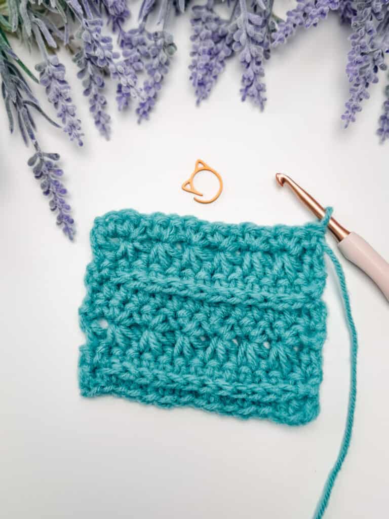 A teal crocheted swatch, a crochet hook, and an orange stitch marker are arranged on a white surface with lavender flowers in the background.
