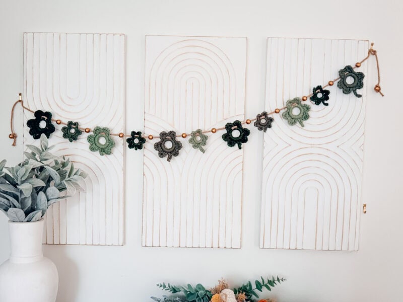 Three white wall panels with geometric patterns, a crochet flower garland hanging across them, and greenery in a white vase and below the panels.