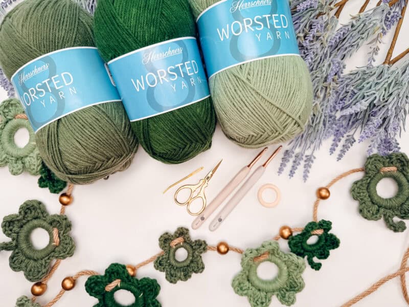 Three skeins of green worsted yarn, scissors, crochet hooks, tape, faux lavender sprigs, and a green crocheted flower garland arranged on a white surface.