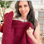 Woman holding a chunky, red Petal Loop Stitch Blanket, smiling at the camera. Text reads: “Cozy Blanket You’ll Actually Use. YOU CAN CROCHET THIS!”.