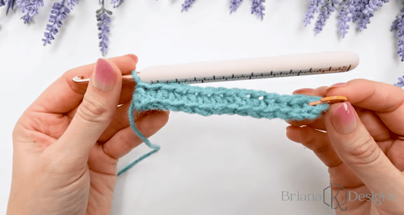 Two hands hold a crochet hook and a length of blue crocheted fabric, with a ruler measuring the width of the stitches.