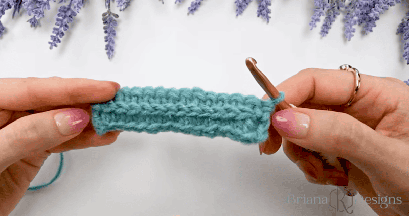 Two hands hold a short, blue crocheted swatch with a crochet hook inserted into one end. Lavender sprigs are visible in the background.