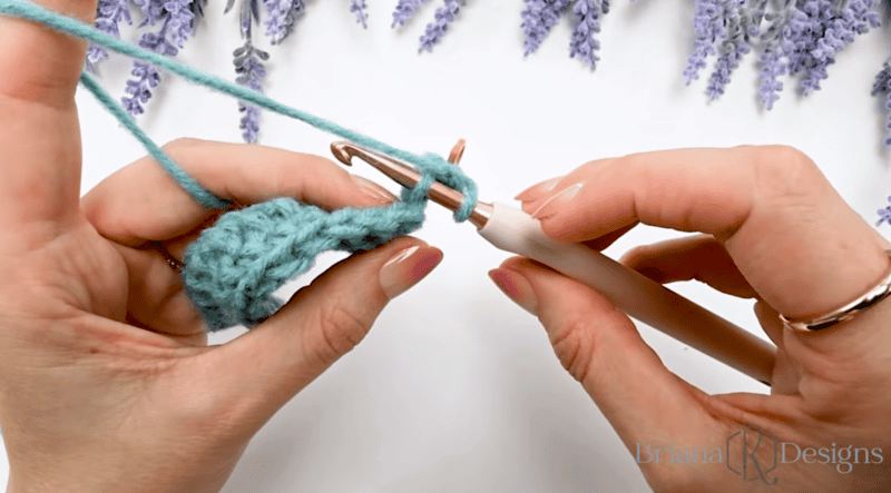 Close-up of hands crocheting with light teal yarn and a crochet hook against a white background with lavender flowers at the top.