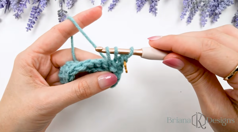 A person crochets with a blue yarn and a white crochet hook, holding a partially completed stitch; lavender flowers are arranged at the top of the frame.