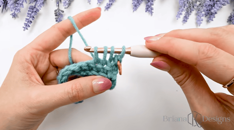 Close-up of hands crocheting with a turquoise yarn and a white crochet hook; lavender flowers are in the background.