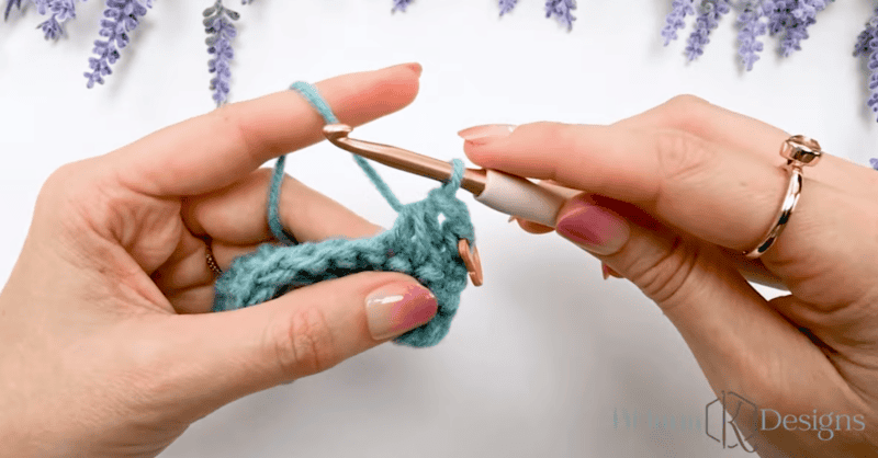 Hands holding a crochet hook and blue yarn, working on a crochet project, with lavender sprigs in the background.