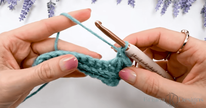 Close-up of hands holding blue yarn and a crochet hook while working on a crochet project, with lavender flowers in the background.