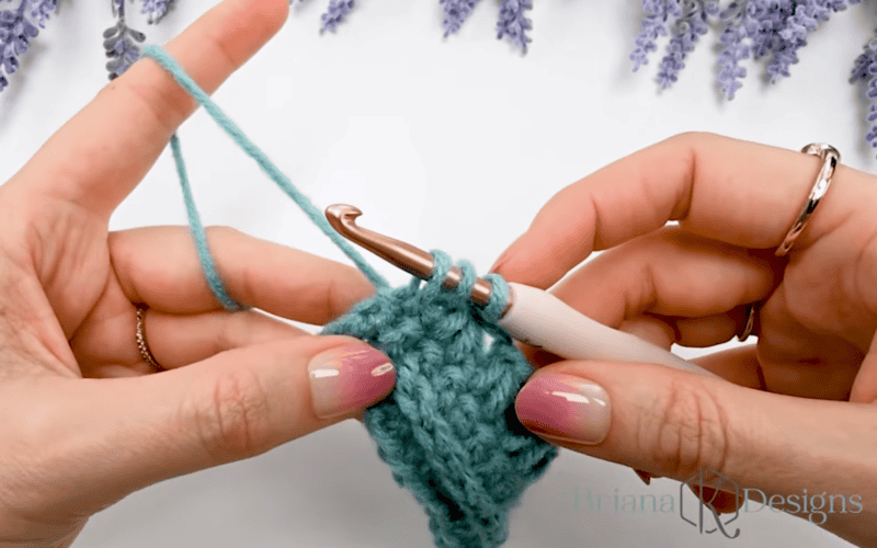 Close-up of hands crocheting with teal yarn and a white crochet hook, working on a textured piece; lavender decorations are visible in the background.