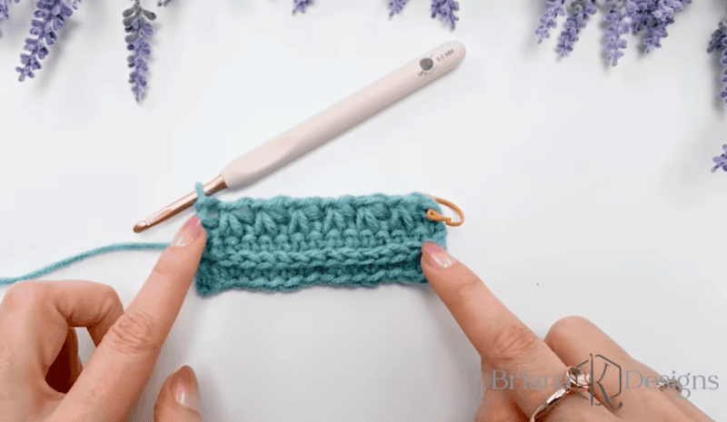 Close-up of hands holding a piece of teal crochet with a gold stitch marker, next to a crochet hook and sprigs of artificial lavender.