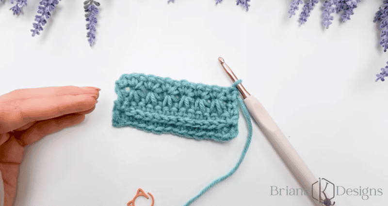 A hand next to a crochet hook and a small blue crocheted swatch, with purple flowers and the Briana K Designs logo in the corners.