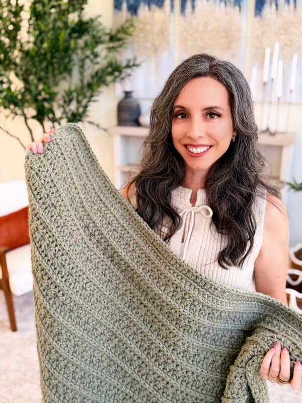 A woman with long gray hair smiles while holding up a textured green crocheted petal loop stitch blanket indoors, reminiscent of the Petal Loop Stitch Trivet design.