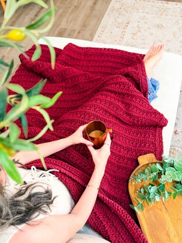 A person sits on a couch with a red knitted blanket, holding a cup of tea or coffee, with a Petal Loop Stitch Trivet on the wooden tray and a green leafy branch nearby.