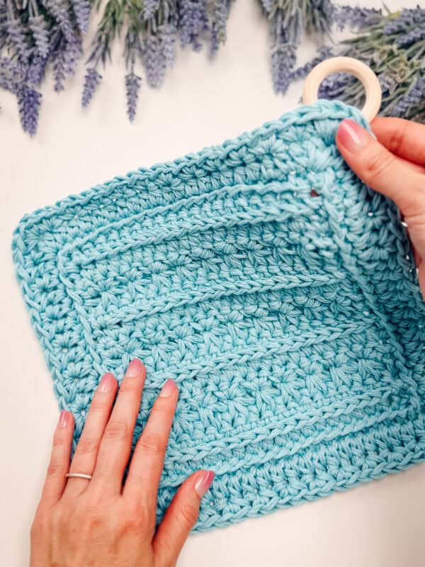 Two hands hold a blue crocheted square with textured patterns and a wooden ring in one corner, placed on a white surface with lavender flowers at the top.
