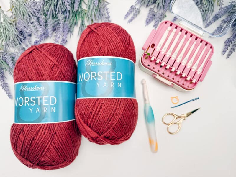 Two skeins of red worsted yarn, a crochet hook, embroidery scissors, stitch marker, hairpin lace loom, and a bobby pin rest on a white surface with lavender sprigs&mdash;perfect essentials for crafting a Petal Loop Stitch Trivet.