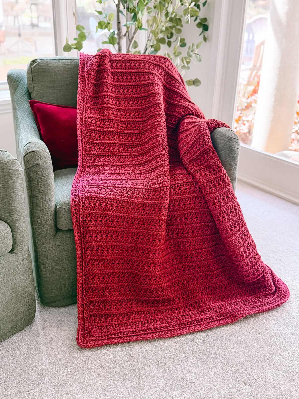 A textured red Petal Loop Stitch Blanket is draped over a green armchair with a matching red pillow, next to a window and a potted plant.
