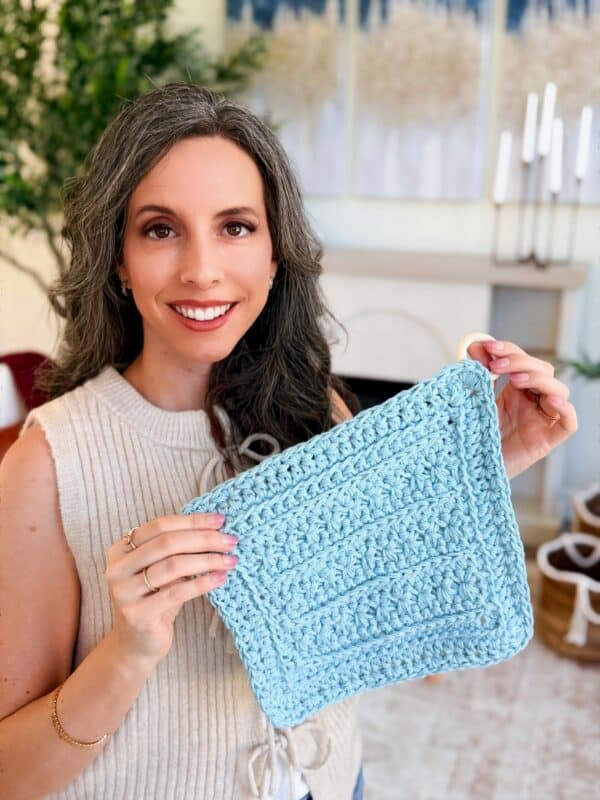 A woman with long gray hair smiles while holding up a light blue crocheted square in a bright, modern room.