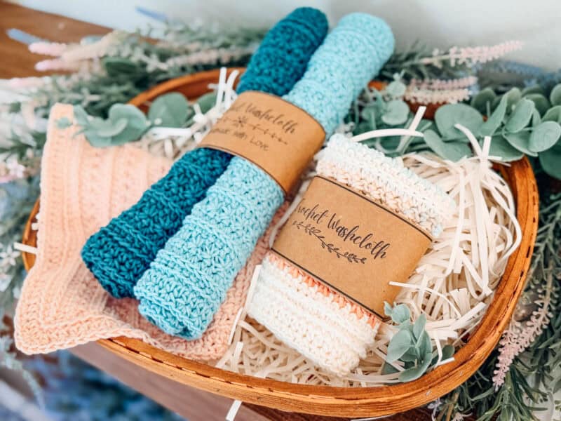 A wicker basket filled with crocheted petal loop stitch washcloths in blue, peach, and cream tones, some rolled and others folded, surrounded by decorative greenery and faux straw.