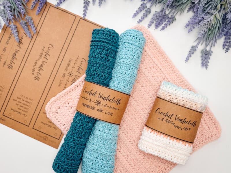 Two rolled crochet washcloths with labels and one folded washcloth are displayed on a white surface, with lavender sprigs and brown product tags in the background.