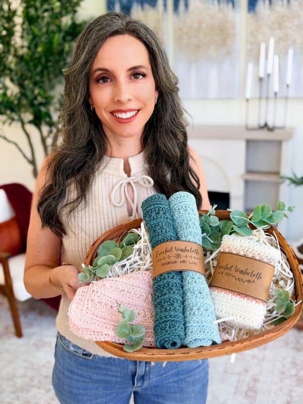A woman holding a wooden tray with four rolled crochet petal loop stitch washcloths in different colors, decorated with greenery, inside a cozy living room.