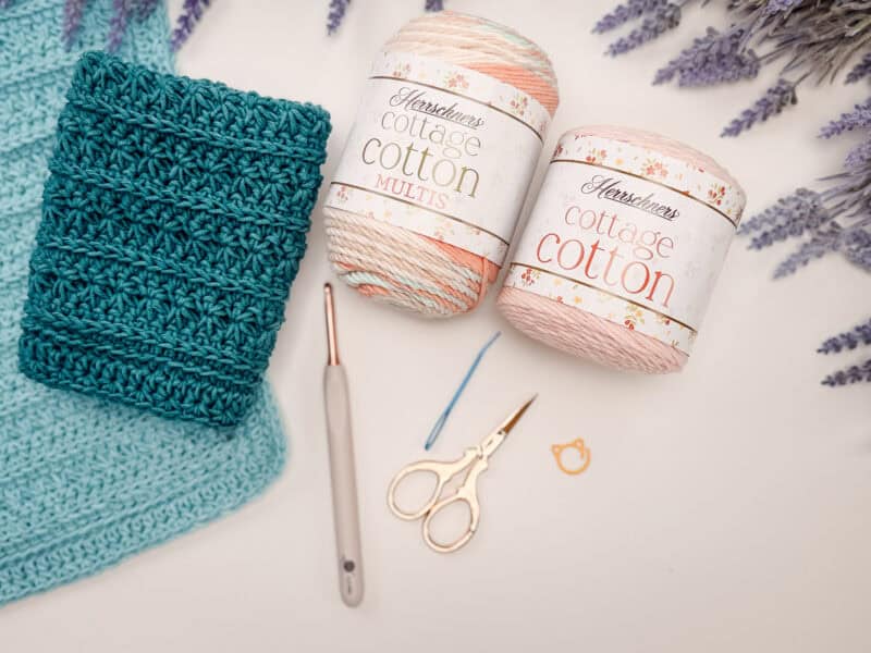 Two skeins of cotton yarn, a pair of scissors, a crochet hook, a needle, a stitch marker, and a teal crocheted petal loop stitch washcloth are arranged on a white surface with lavender sprigs nearby.