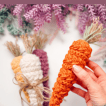 A hand holds a crocheted carrot beside two more crocheted carrots, one orange and one cream and purple, on a white surface with purple foliage above.