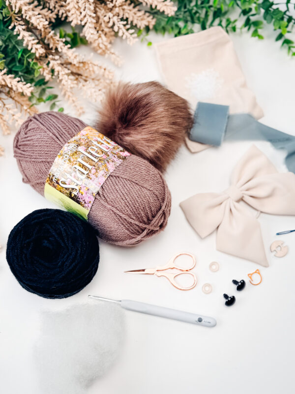 A flat lay of crafting supplies including yarn, a faux fur pompom, a bow, scissors, crochet hook, stitch markers, eyes, fiberfill, and greenery in the background.