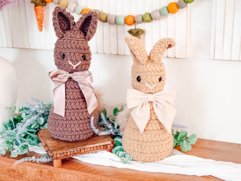 Two crocheted bunny dolls with large bows, one brown and one tan, are displayed on a wooden surface with greenery and decorative garland in the background.