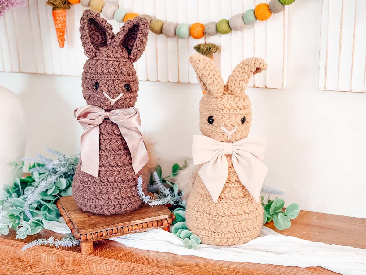 Two crocheted bunny dolls with large bows, one brown and one tan, are displayed on a wooden surface with greenery and decorative garland in the background.