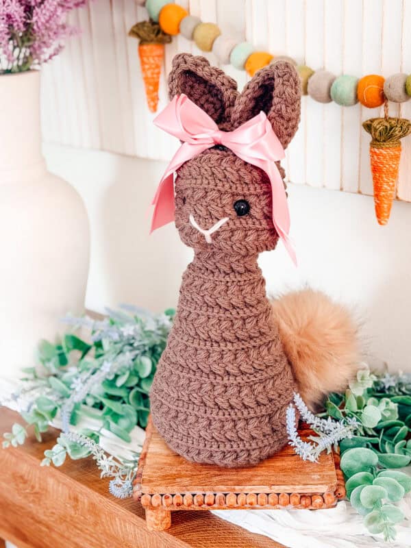 A crocheted brown bunny made with the Wheat Stitch Bunny Crochet Pattern, featuring a pink ribbon and fluffy tail, sits on a wooden surface, surrounded by artificial greenery and decorative carrots.