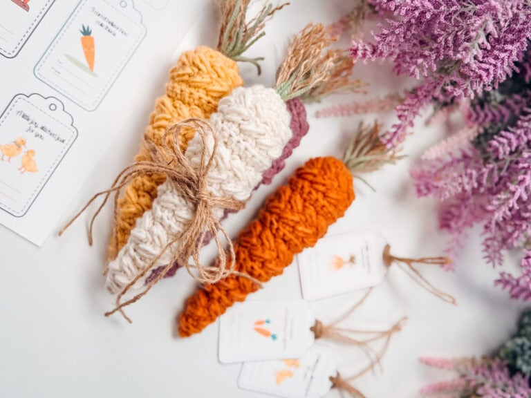 Three crocheted carrots, one tied with twine, are placed on a white surface next to purple flowers and illustrated tags featuring carrot drawings.
