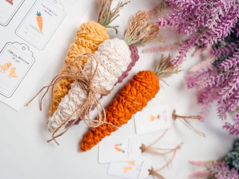 Three crocheted carrots, one tied with twine, are placed on a white surface next to purple flowers and illustrated tags featuring carrot drawings.