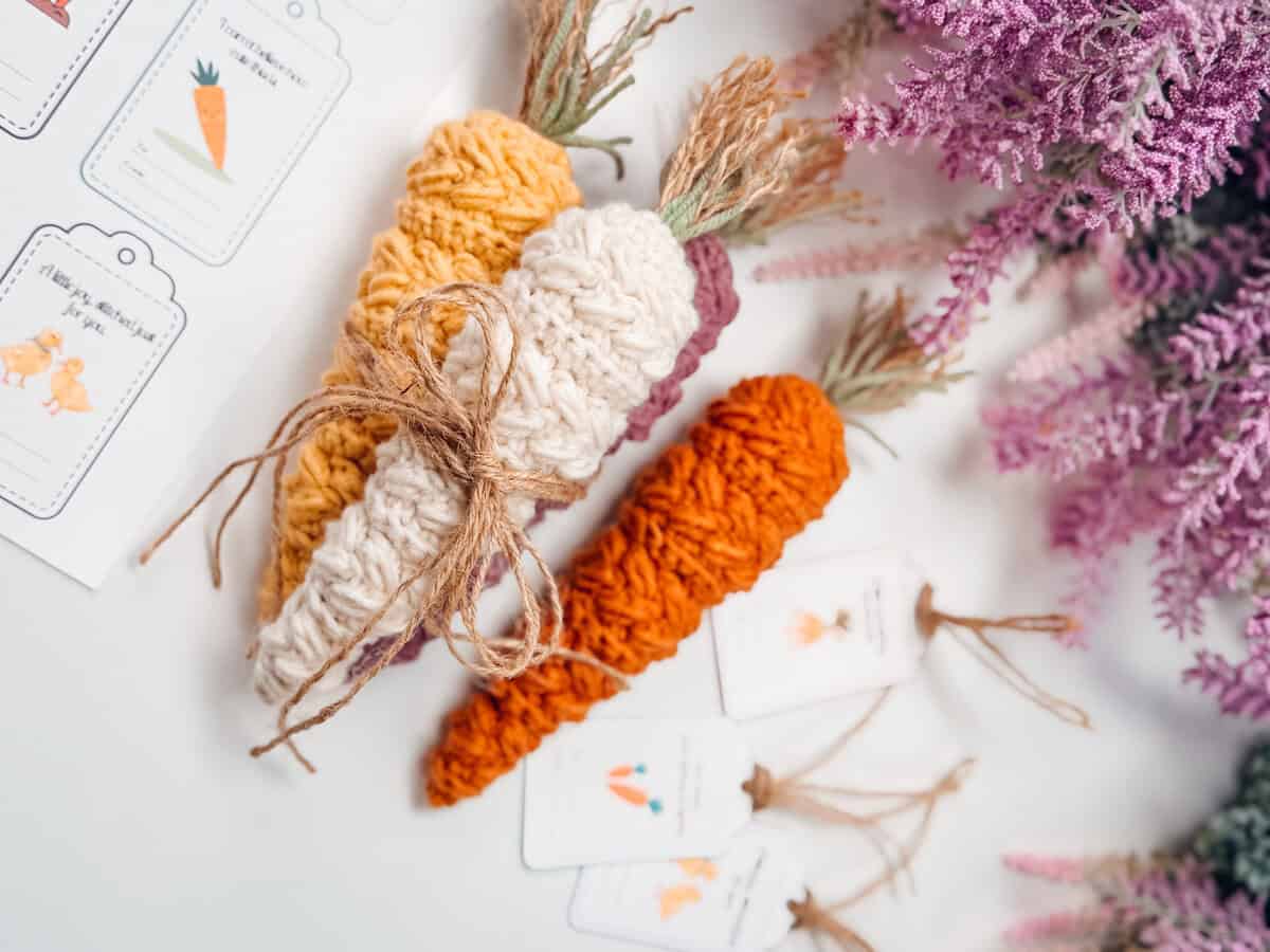 Three crocheted carrots, one tied with twine, are placed on a white surface next to purple flowers and illustrated tags featuring carrot drawings.
