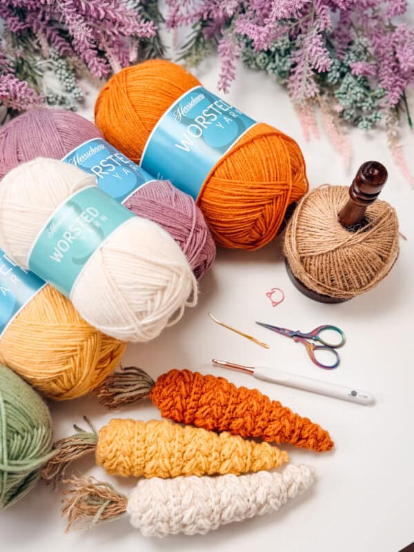 Skeins of yarn, crocheted carrot shapes, scissors, a crochet hook, and other crochet tools are arranged on a table with purple flowers in the background.