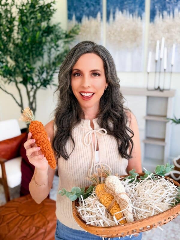 A woman with long dark hair holds a handmade orange carrot crafted from a Wheat Stitch carrot crochet pattern, along with a basket filled with decorative carrots and shredded paper, standing in a bright, modern room.