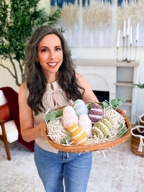 A woman with long gray hair holds a basket filled with colorful eggs made from the Wheat Stitch Easter Egg Crochet Pattern, standing in a cozy, decorated living room.