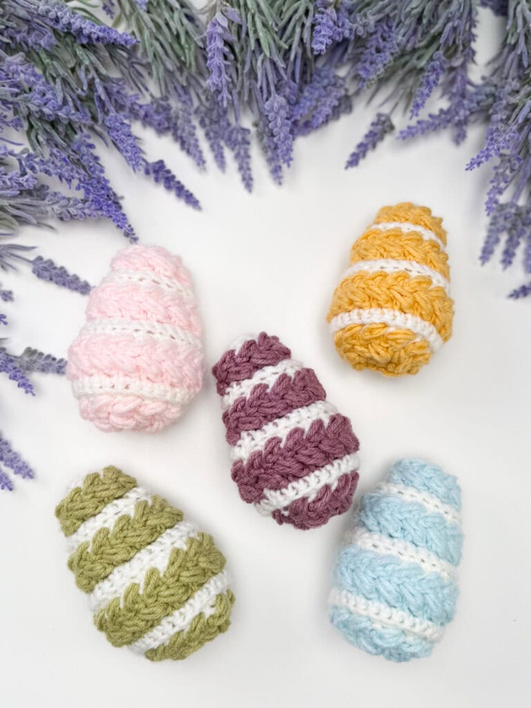 Five crocheted, striped egg shapes in pastel colors arranged in a semicircle on a white surface, with sprigs of lavender flowers at the top of the image.