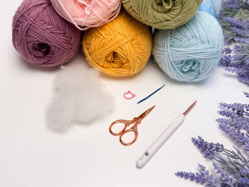 Five balls of yarn, a bundle of stuffing, pink stitch marker, blue yarn needle, gold scissors, and a white crochet hook on a white surface with lavender sprigs.