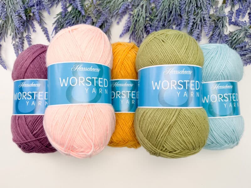 Five skeins of Herschners worsted yarn in purple, pink, yellow, green, and light blue are arranged side by side with lavender flowers in the background.