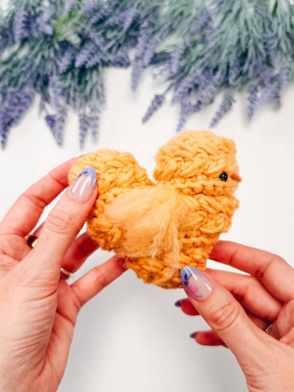 Hands holding a small, yellow, crocheted chick with a black bead eye; lavender flowers blurred in the background.