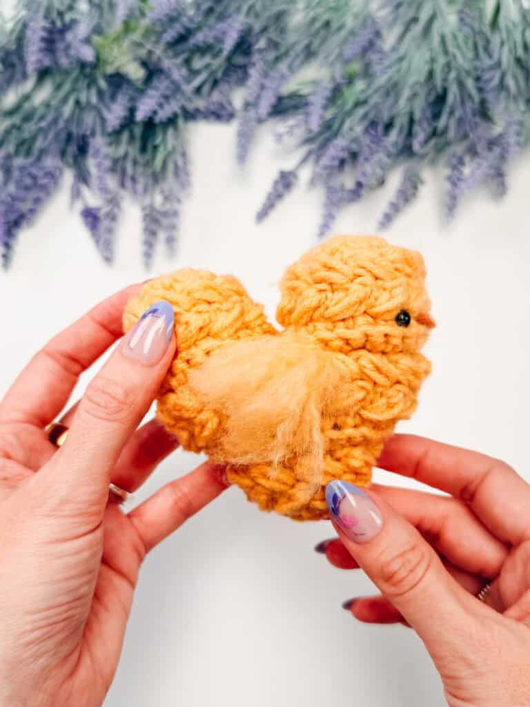 Hands holding a small, yellow, crocheted chick with a black bead eye; lavender flowers blurred in the background.