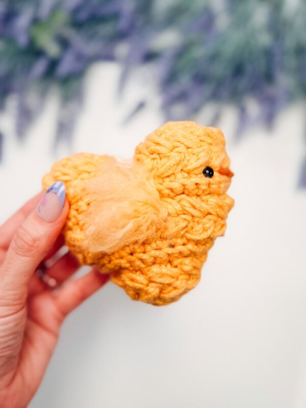 A hand holding a small, yellow, crocheted chick from the wheat stitch chick crochet pattern with a black eye and orange beak, against a blurred background.