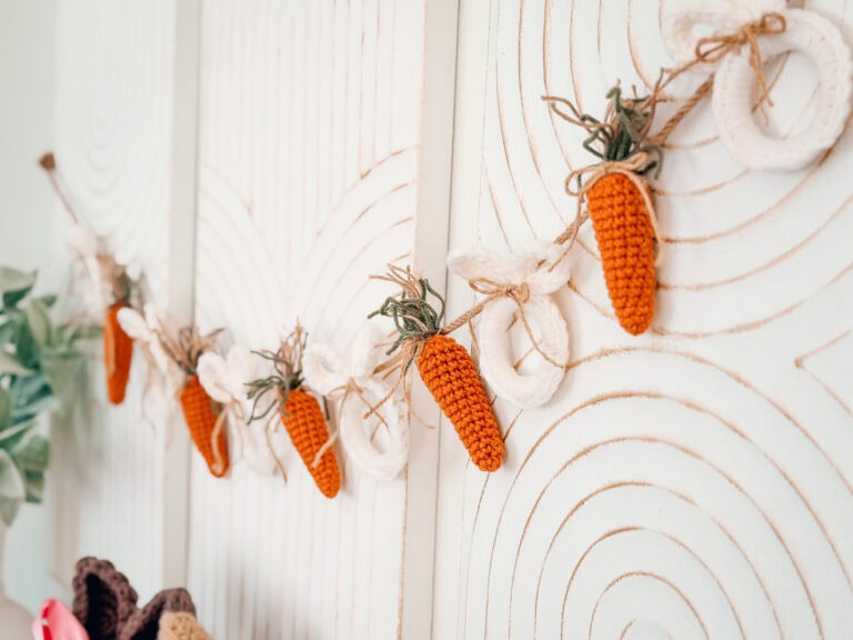 A decorative garland with crocheted orange carrots and white rings hangs on a white textured wall.