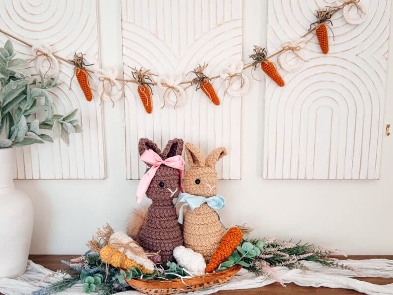 Two crocheted bunnies with ribbons sit among artificial greenery and carrots, featuring a mini crochet carrot pattern garland hanging on a white textured wall in the background.