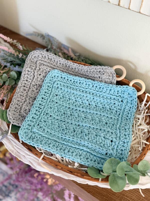 Two crocheted pot holders, one gray and one light blue, crafted with the Petal Loop Stitch Trivet technique, are displayed in a basket with decorative greenery and shredded paper.