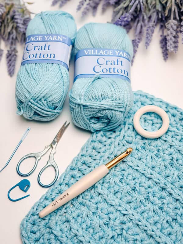 Two skeins of blue craft cotton yarn, crocheted fabric, scissors, crochet hook, stitch marker, and a wooden ring arranged on a white surface with lavender flowers above.