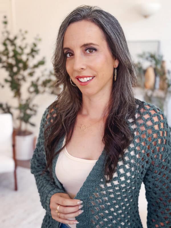 A woman with long, wavy gray hair wears a green crochet cardigan pattern over a white top and gold jewelry, standing indoors with plants and light furniture in the background.